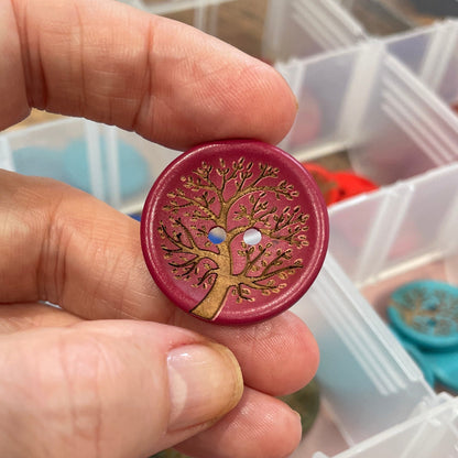 Hand holding a decorative button with intricate design, surrounded by various buttons in a clear container.