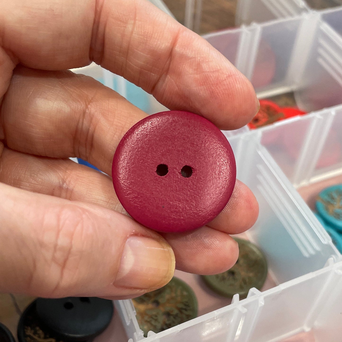 Hand holding a red button with a collection of buttons in clear containers in the background.