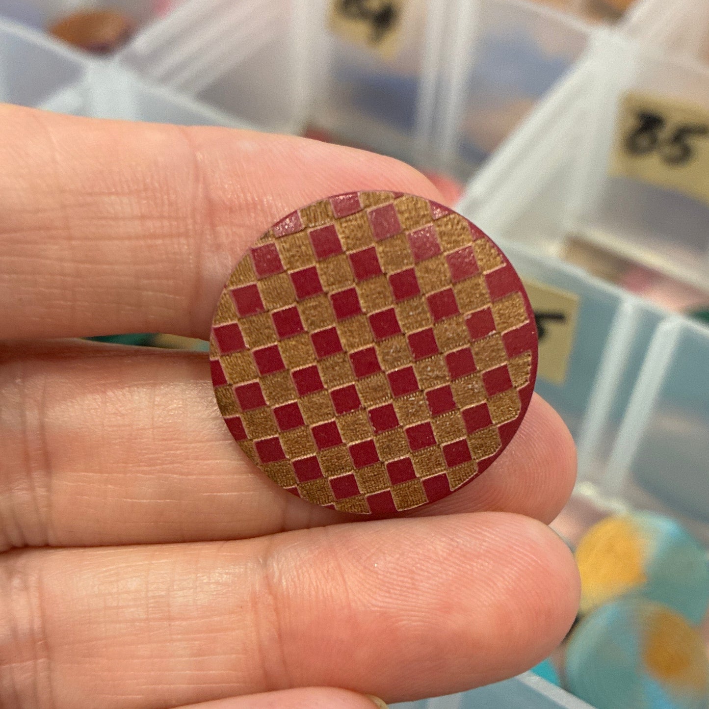 Hand holding a red and gold woven button with a container of buttons in the background