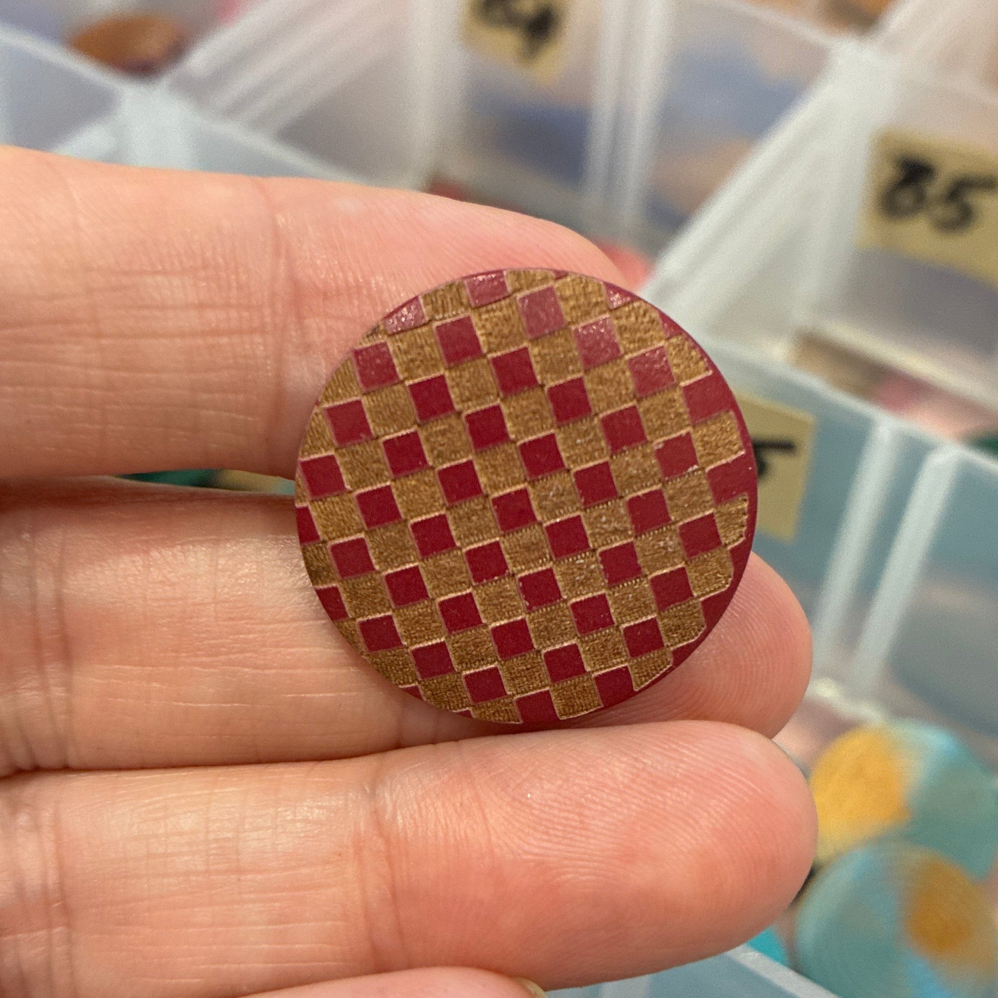 Hand holding a red and gold woven button with a container of buttons in the background