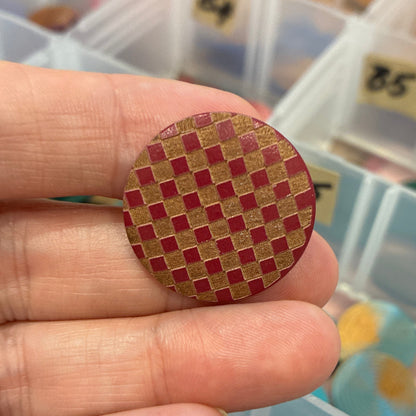 Hand holding a red and gold woven button with a container of buttons in the background