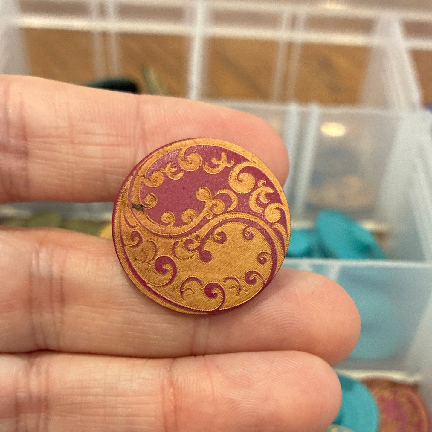 Close-up of a hand holding a decorative button with a blurred background of a storage box with buttons.