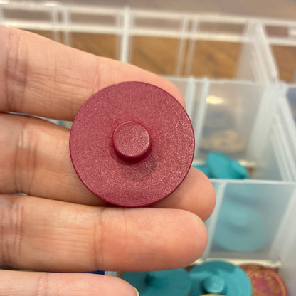 Hand holding a round pink button with a container of colorful objects in the background