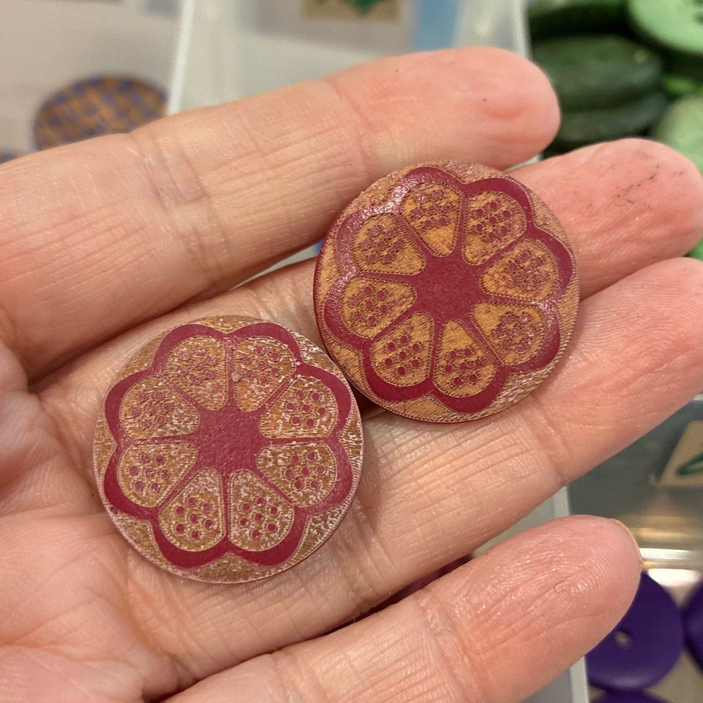 Hand holding two decorative buttons with a container of more buttons in the background.