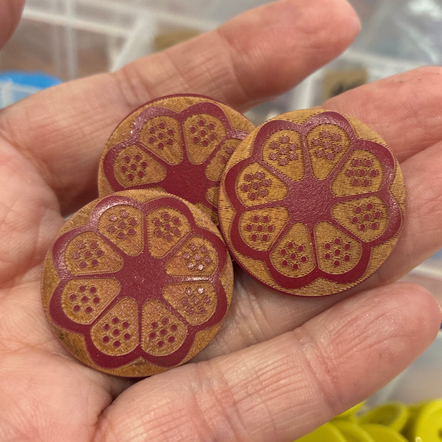 Hand holding two decorative buttons with a floral pattern, with a container of buttons in the background.