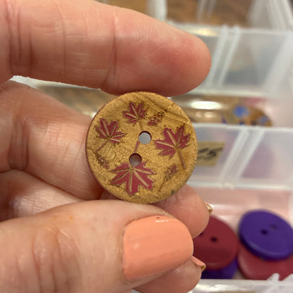 Hand holding a decorative button with floral patterns against a background of button storage containers.
