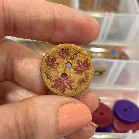 Hand holding a decorative button with floral patterns against a background of button storage containers.