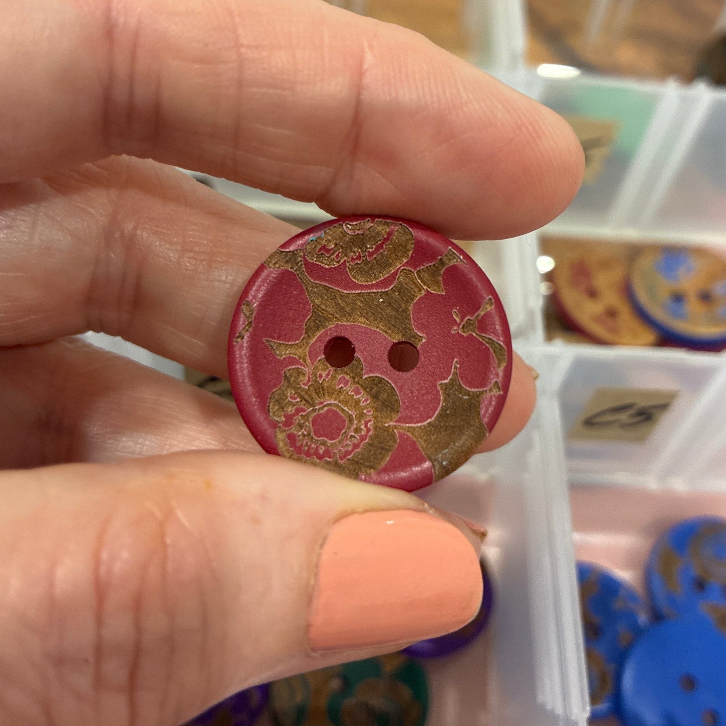 Hand holding a red button with floral design in front of a button collection box.