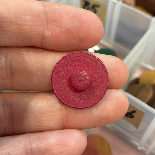 Round red button held between fingers with a container of similar buttons in the background