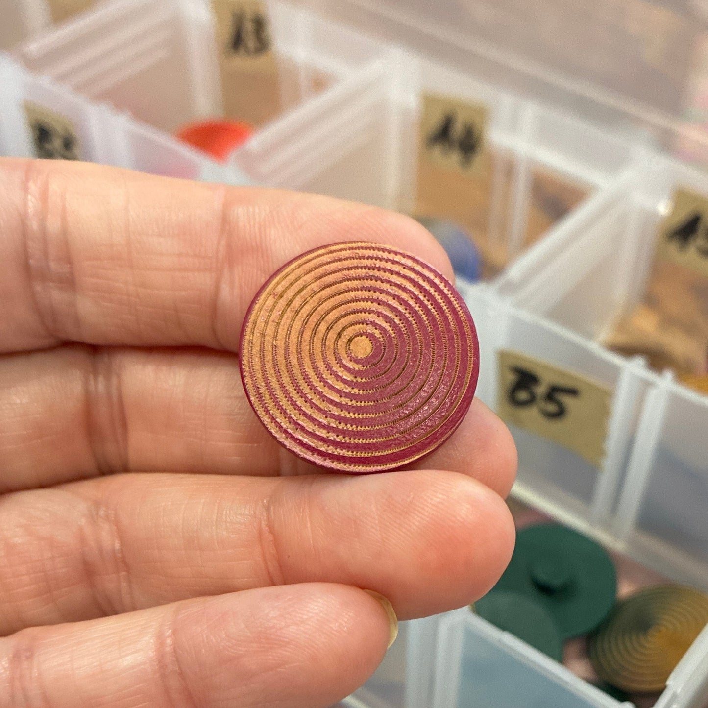 Hand holding a circular button with a detailed pattern, against a background of a clear container with various colored objects.