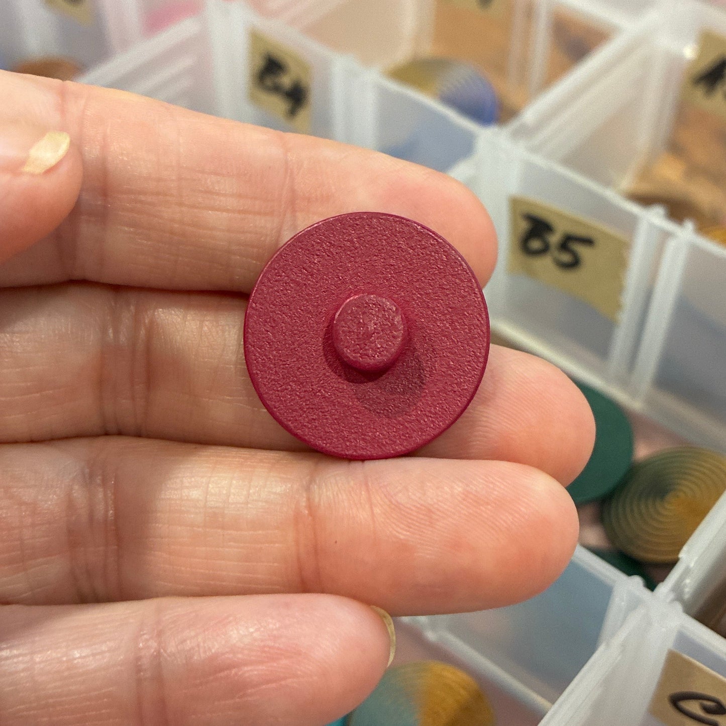 Hand holding a red button with a clear container of assorted buttons in the background