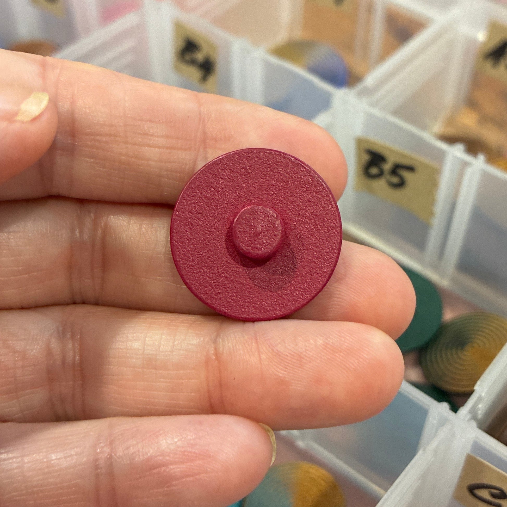 Hand holding a red button with a clear container of assorted buttons in the background