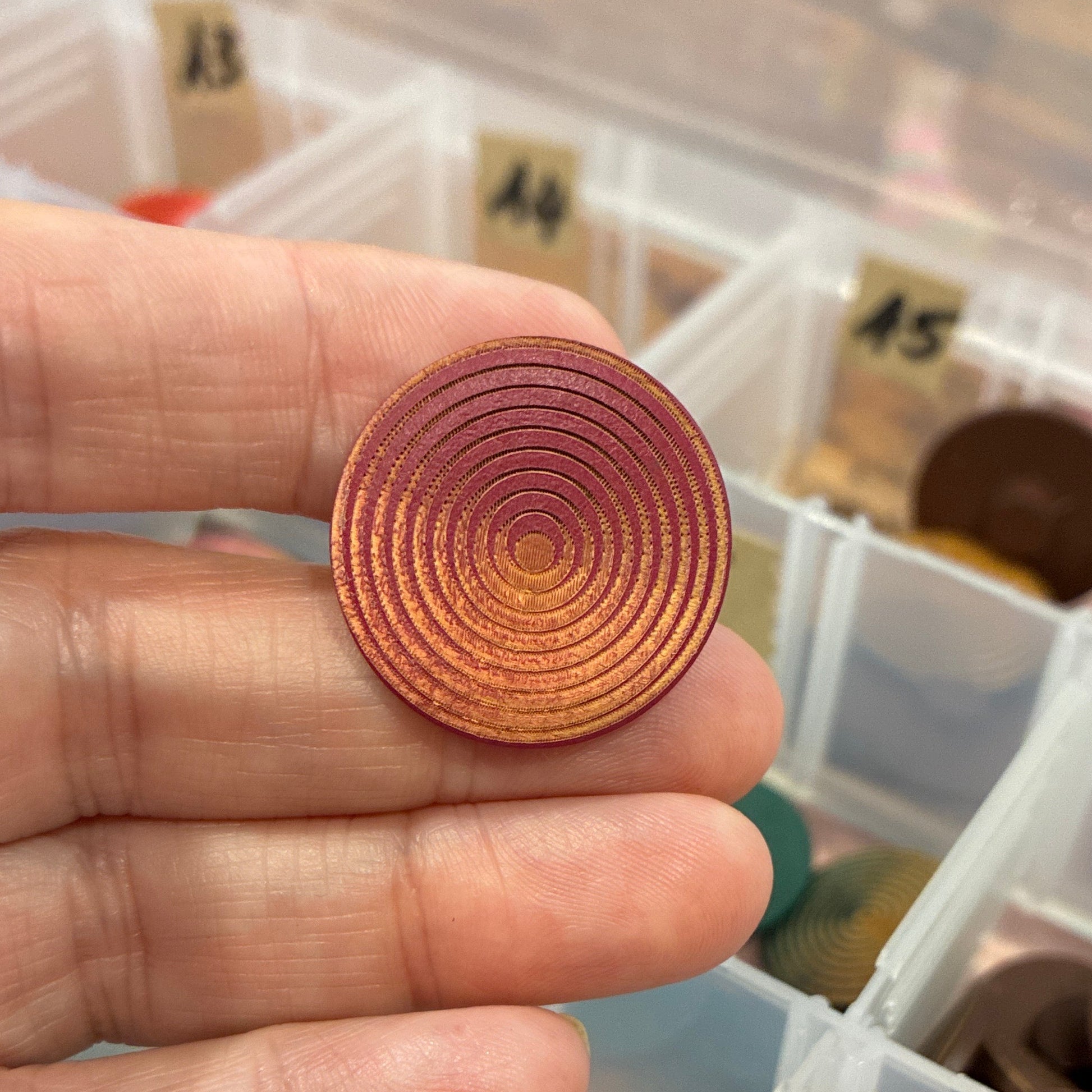 Hand holding a circular copper-colored button with a container of similar buttons in the background.