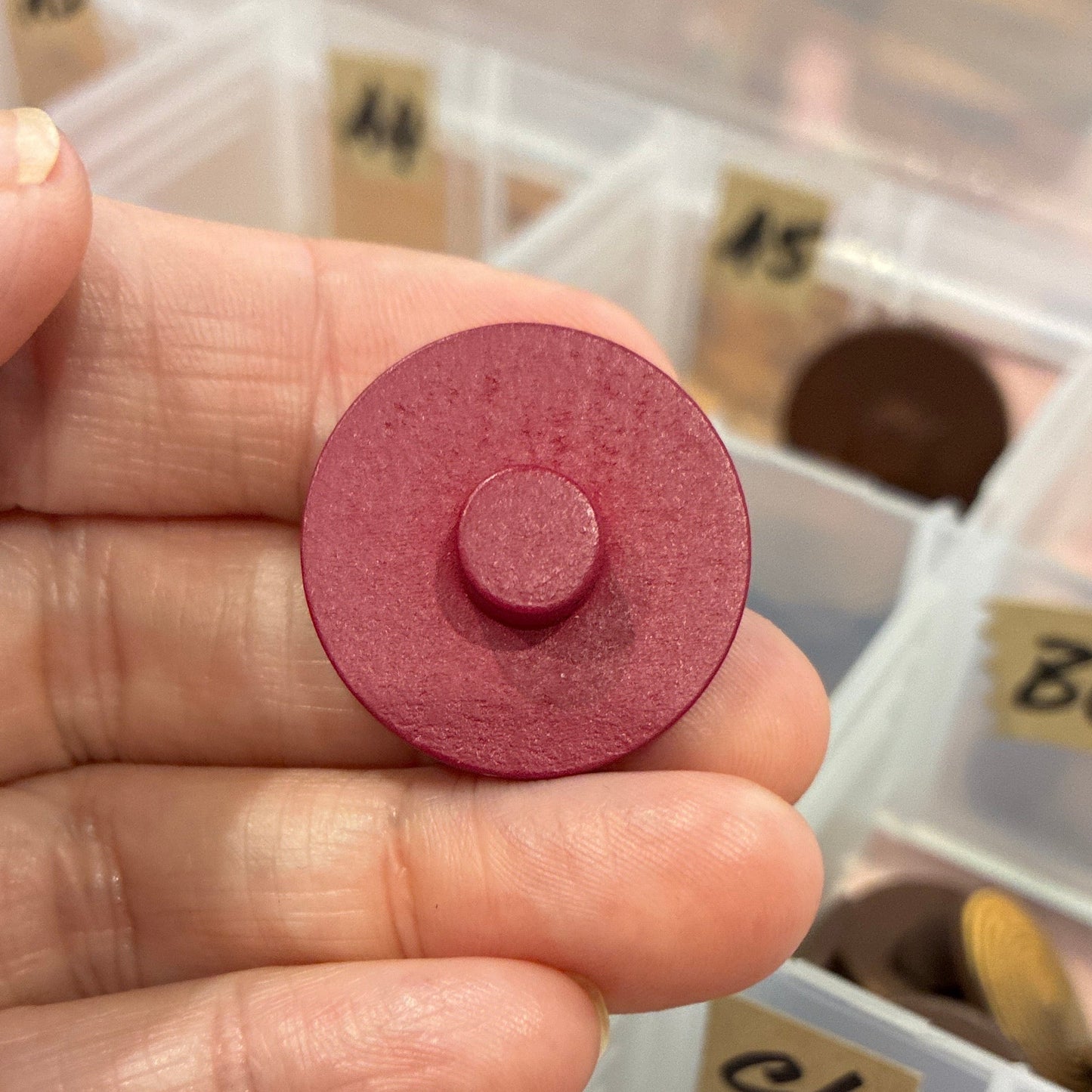 Hand holding a red circular button with a collection of similar objects in a box in the background