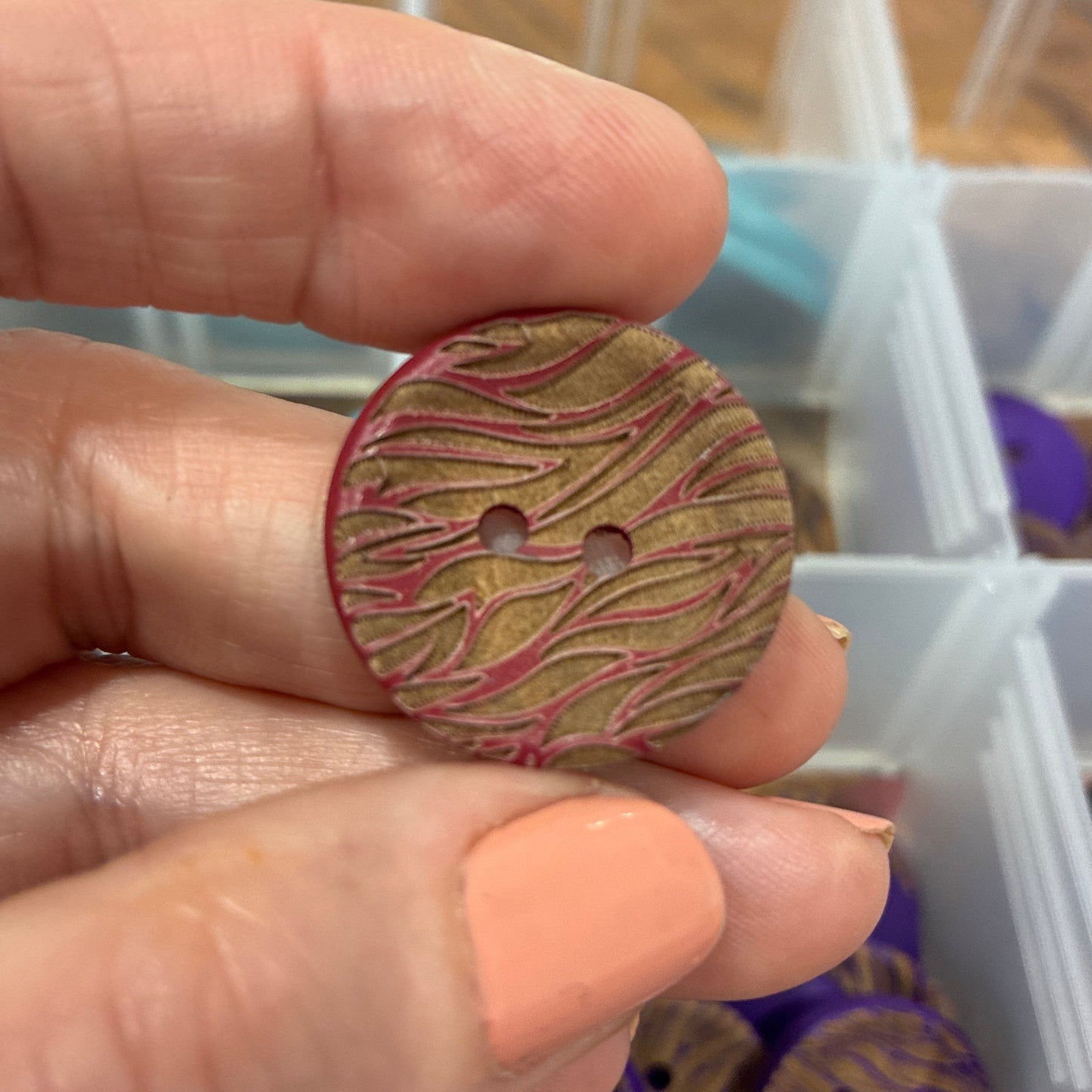 Hand holding a patterned button with a blurred background of a storage box.