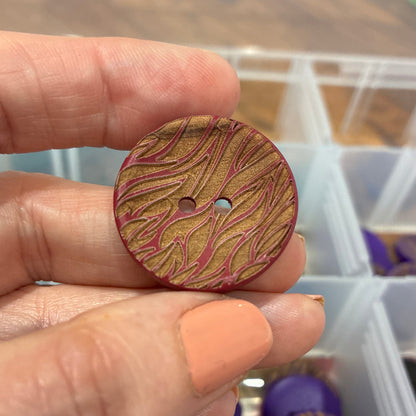Hand holding a wooden button with a pattern, with a container of buttons in the background.