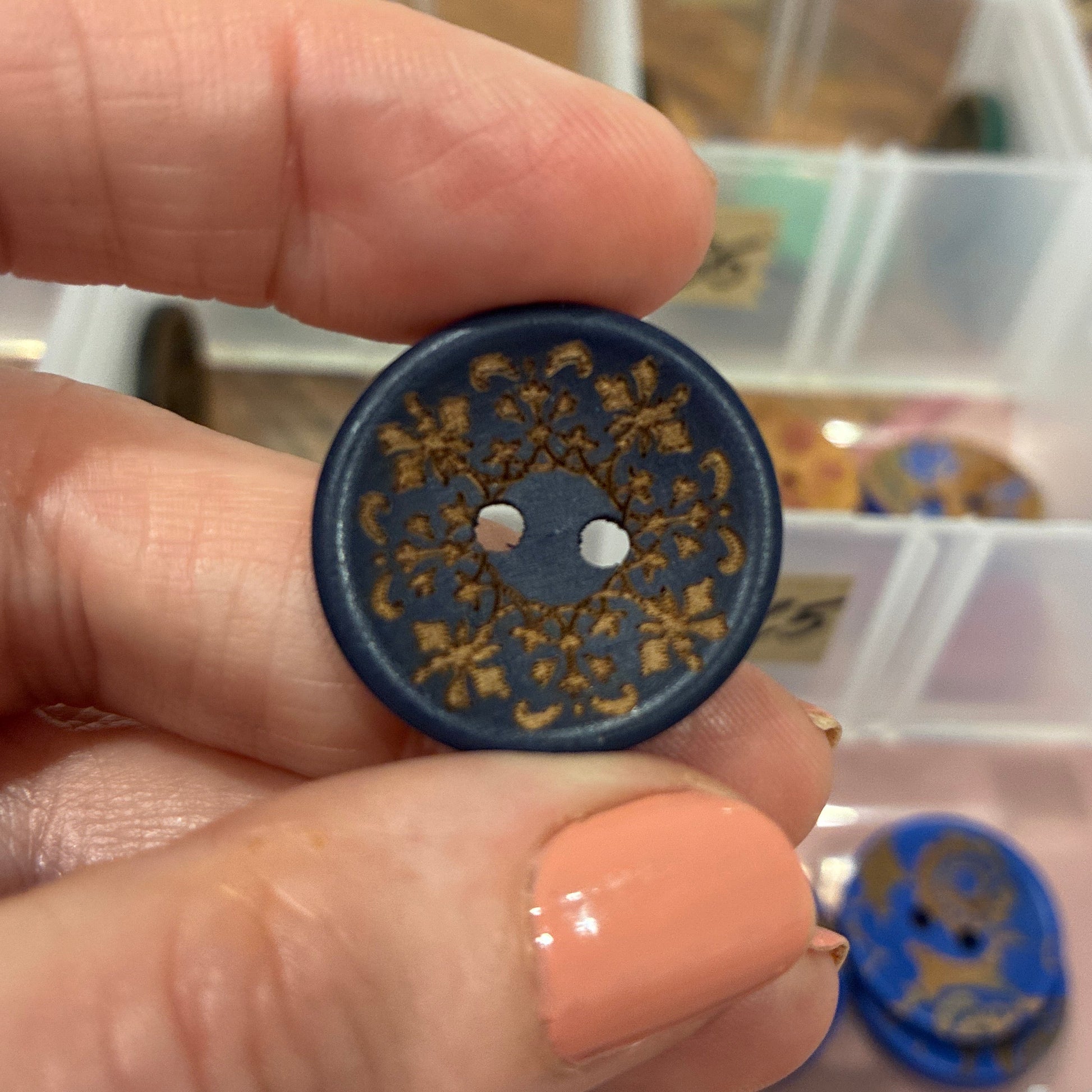 Hand holding a decorative button with intricate designs, blurred background of a storage box.