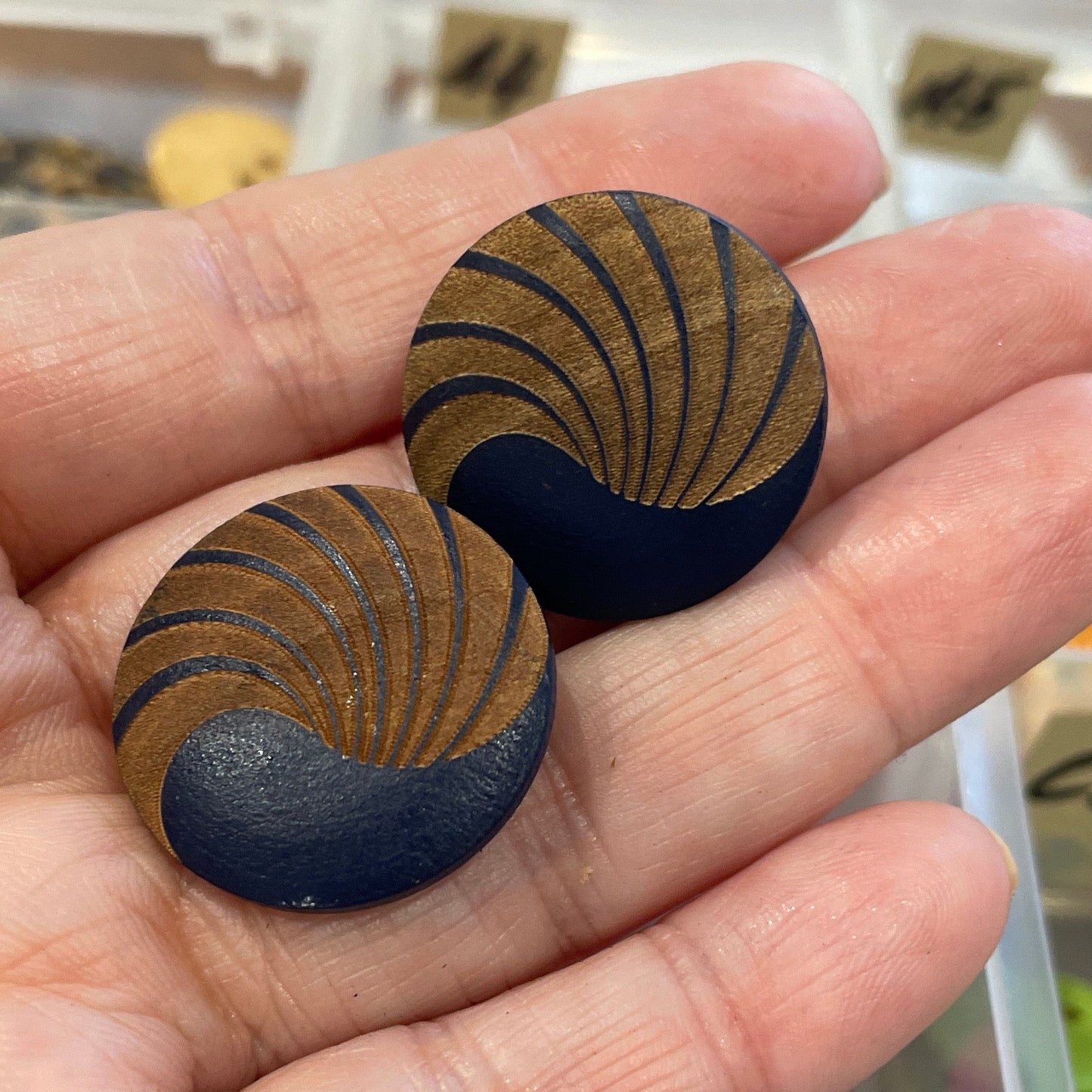 Hand holding two decorative buttons with a background of a button collection in a box.