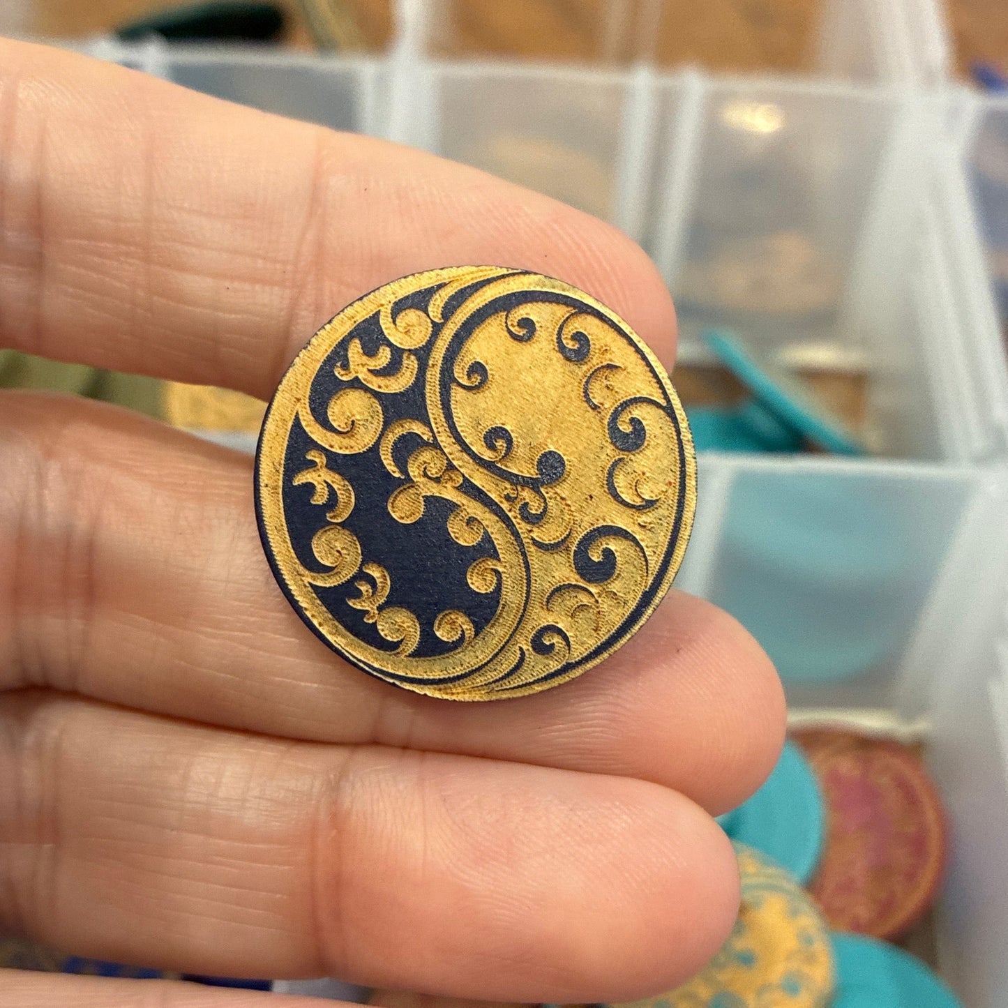Gold and black circular decorative item held between fingers with a colorful background
