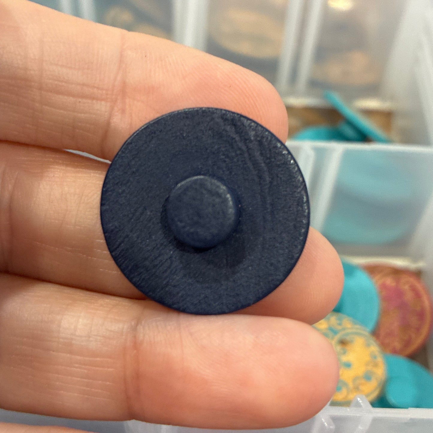 Black round button held between fingers with a blurred background of colorful items in a box.