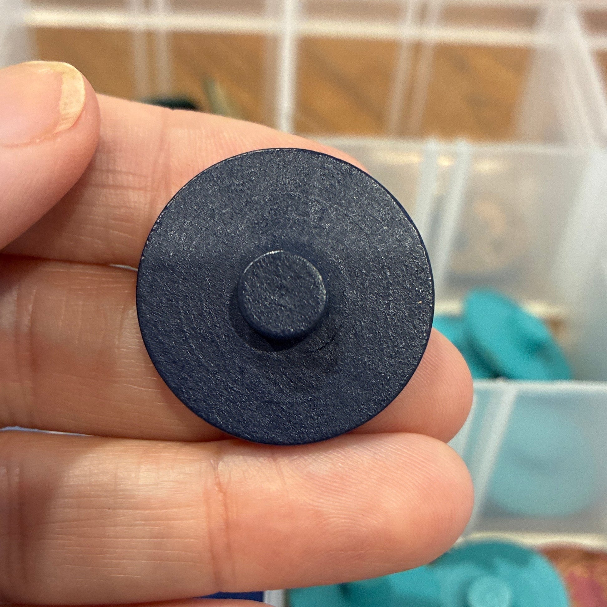 Black circular button held in a hand with a container of colorful buttons in the background