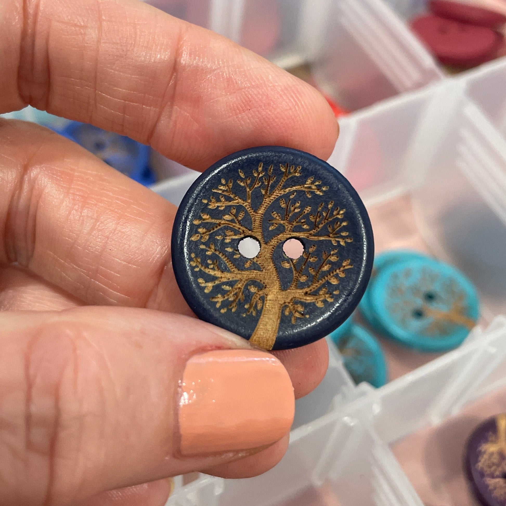 Hand holding a decorative button with intricate design, with a container of buttons in the background.