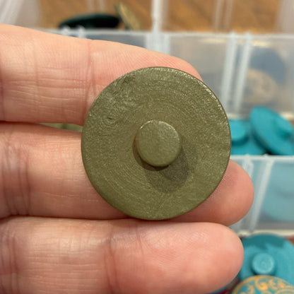 Hand holding a green circular button with a blurred background of colorful items in a container.