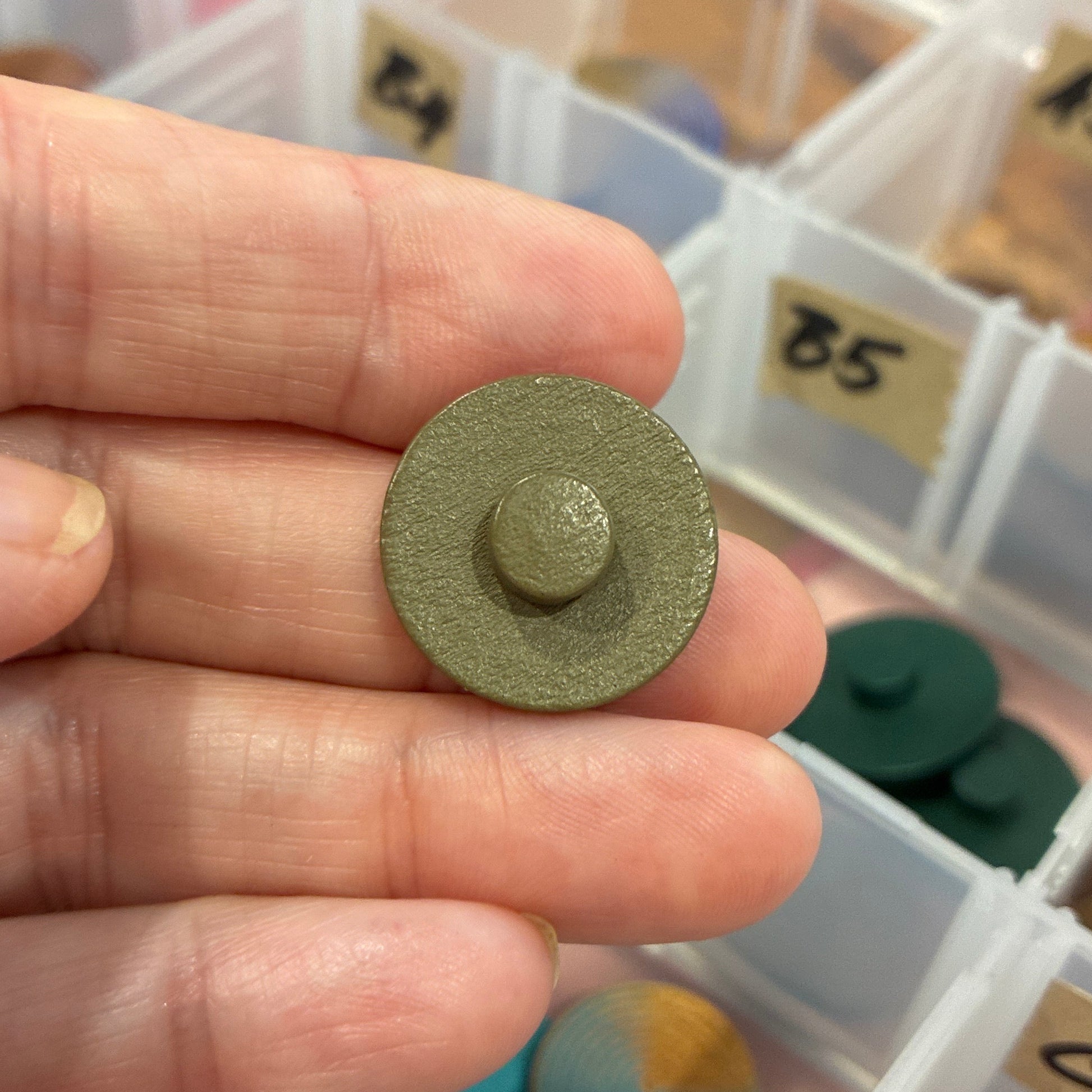 Hand holding a small green button with a colorful assortment of items in the background