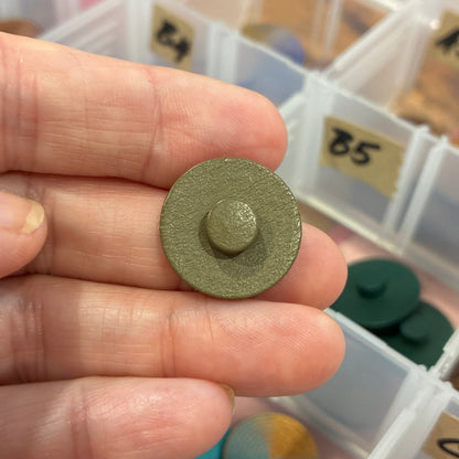 Hand holding a small green button with a colorful assortment of items in the background