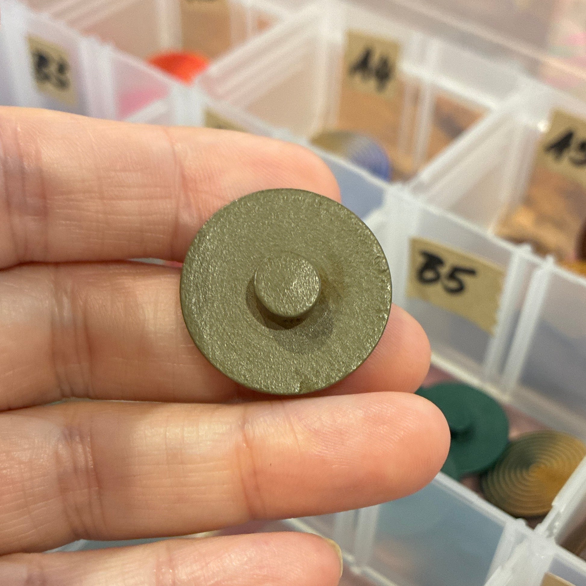 Hand holding a round metal button with a background of color swatches in a box.