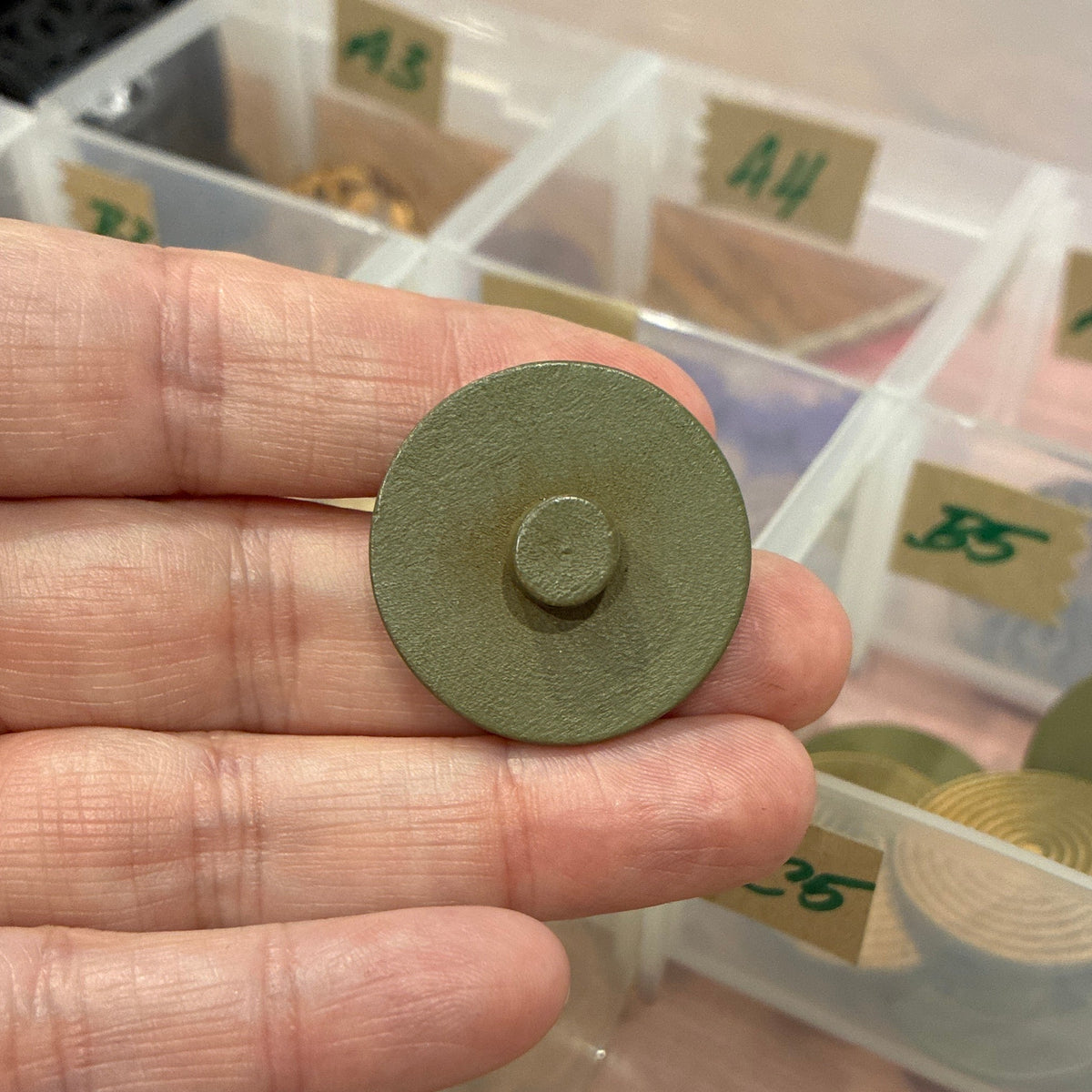 Hand holding a green button with a background of various buttons in a clear container.