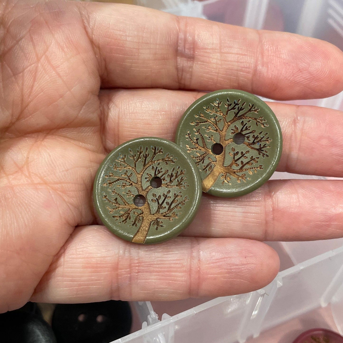 Hand holding two decorative buttons with a container of similar buttons in the background.