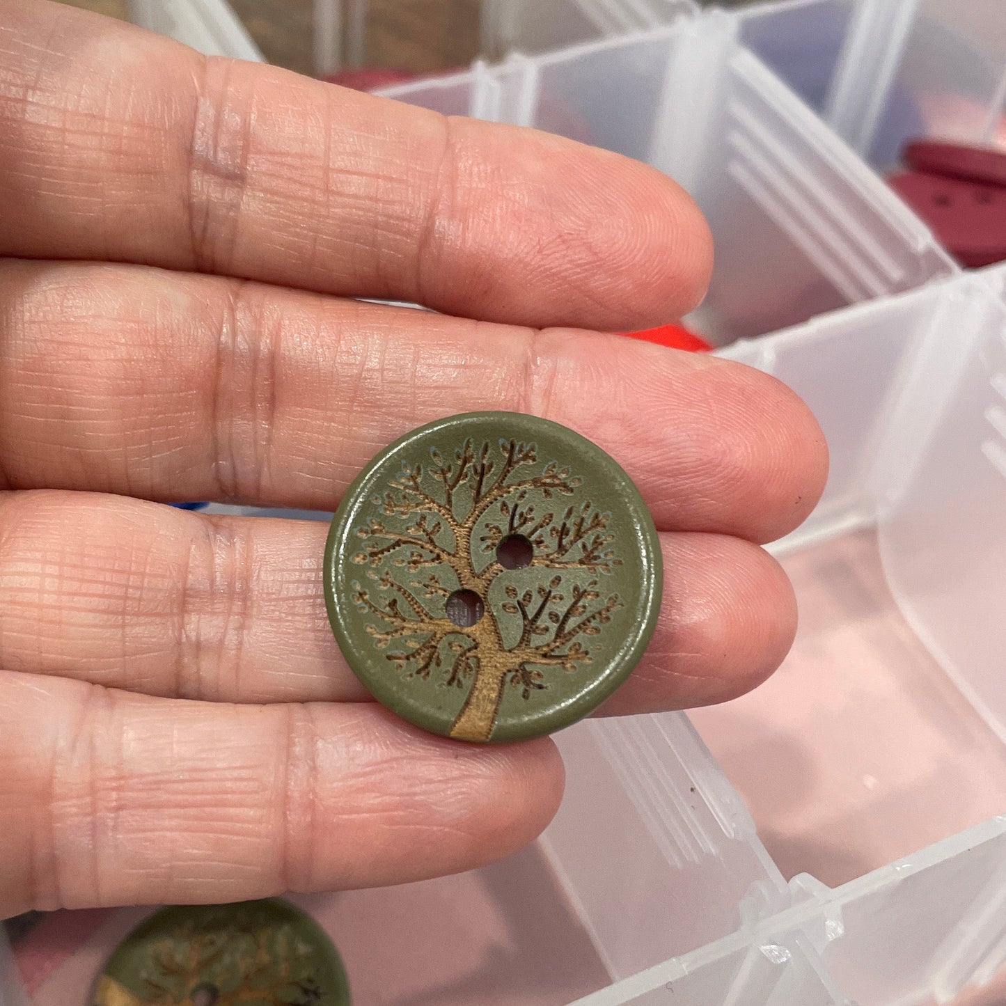 Hand holding a green button with a detailed design, against a background of a clear plastic container with more buttons.