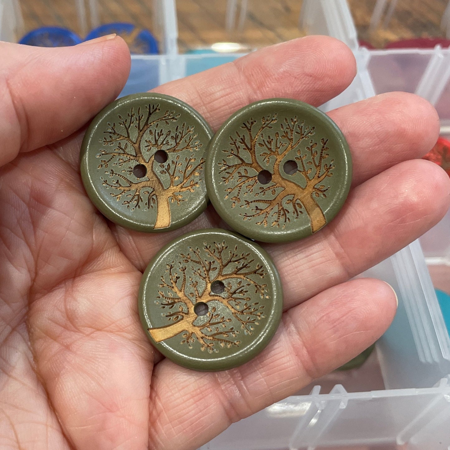 Hand holding three decorative buttons with a background of button storage containers.