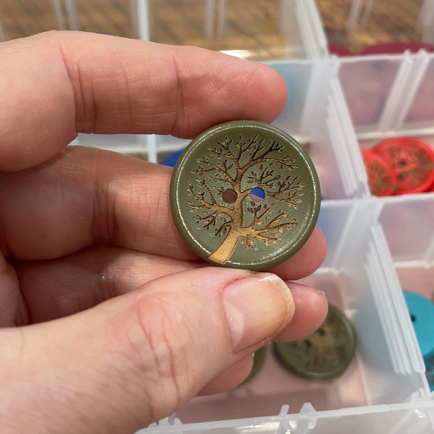 Hand holding a decorative button with a tree design, in front of a container filled with various buttons.