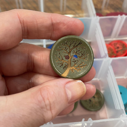 Hand holding a decorative button with a tree design, in front of a container filled with various buttons.