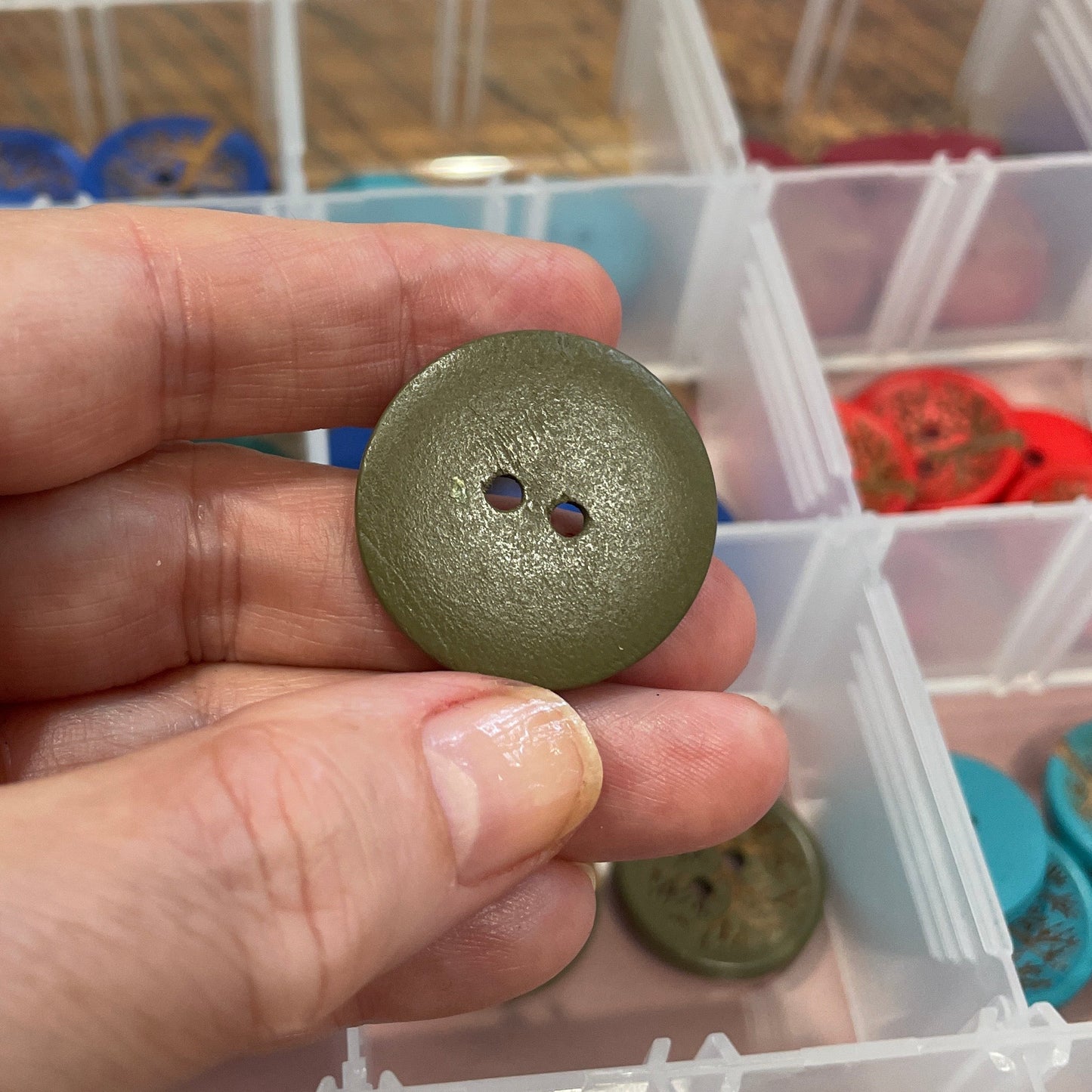 Hand holding a green button with a clear plastic container of buttons in the background