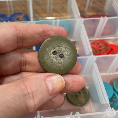 Hand holding a green button with a clear plastic container of buttons in the background