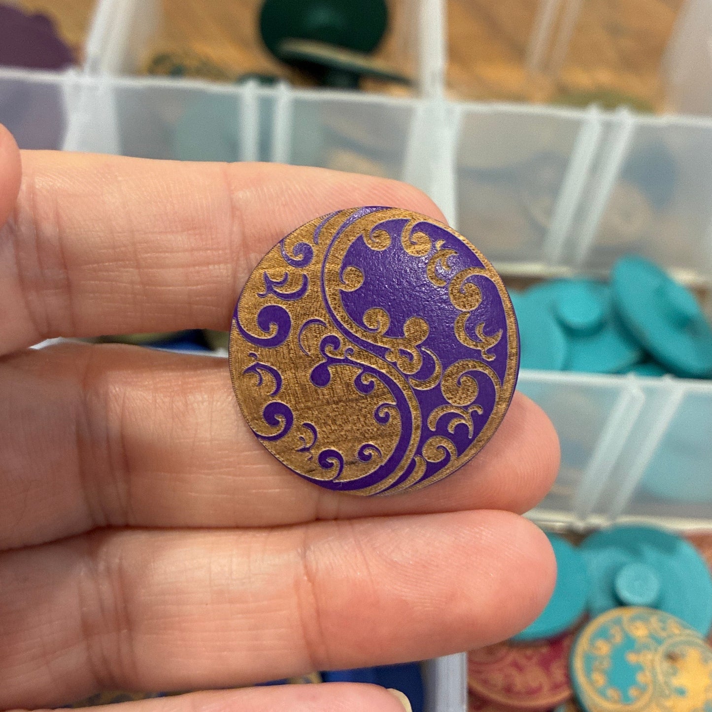 Hand holding a decorative purple and gold button with a blurred container of buttons in the background.