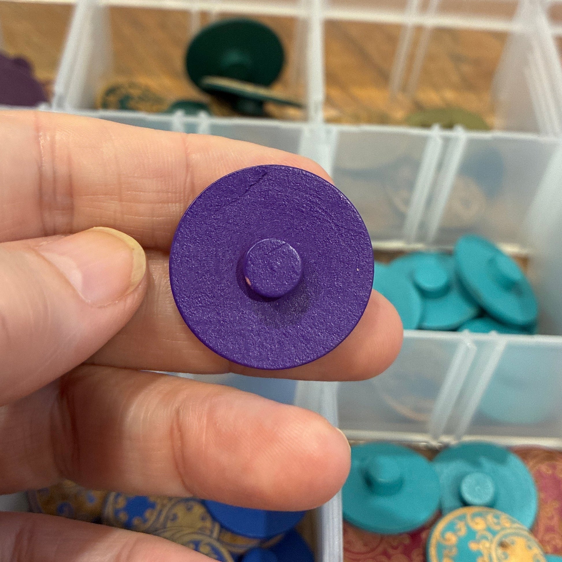 Hand holding a purple button with a container of buttons in the background