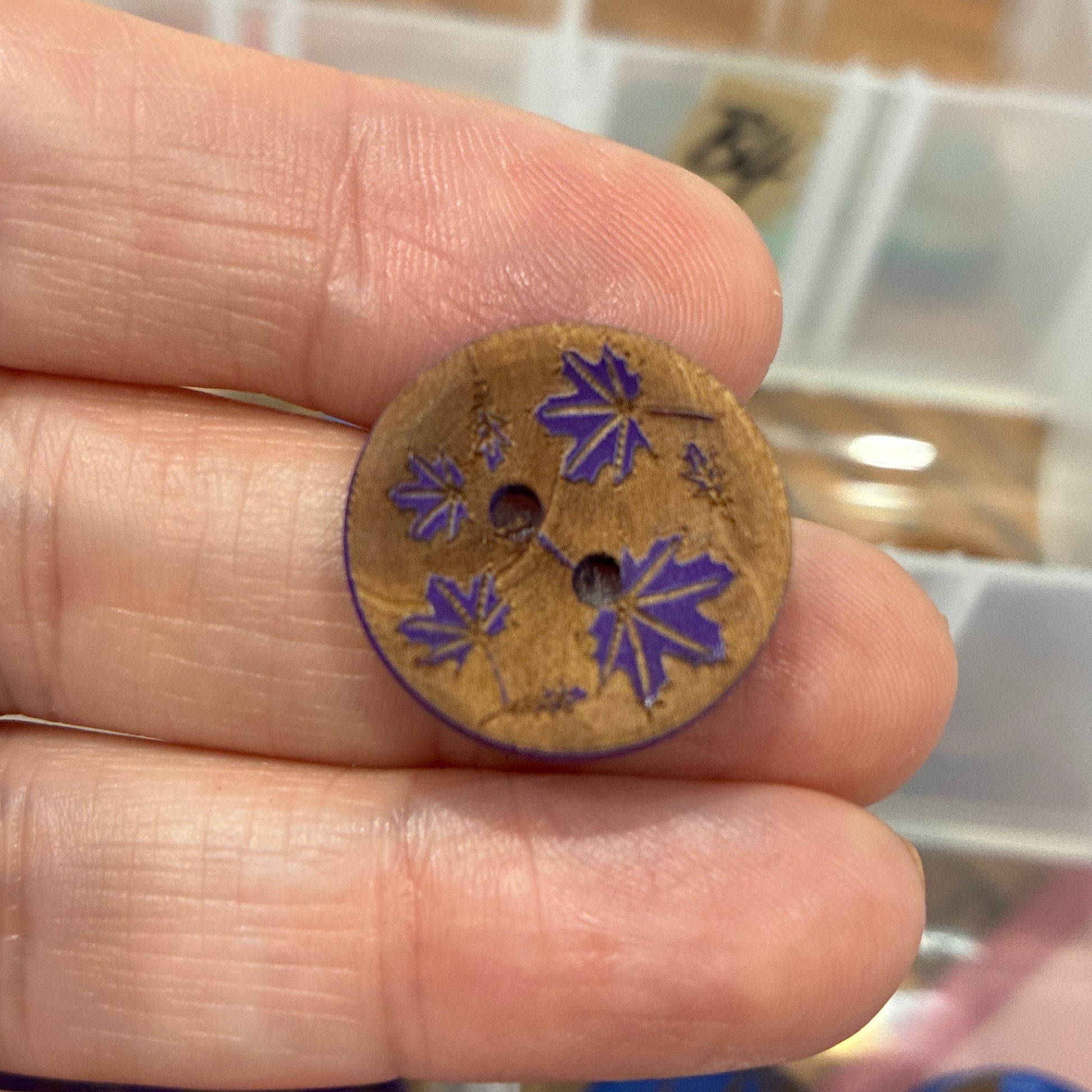 Hand holding a small brown button with purple floral design, blurred background of a storage box.