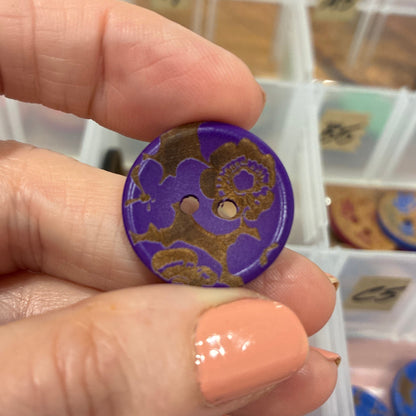 Hand holding a purple button with gold design in front of a container of buttons.