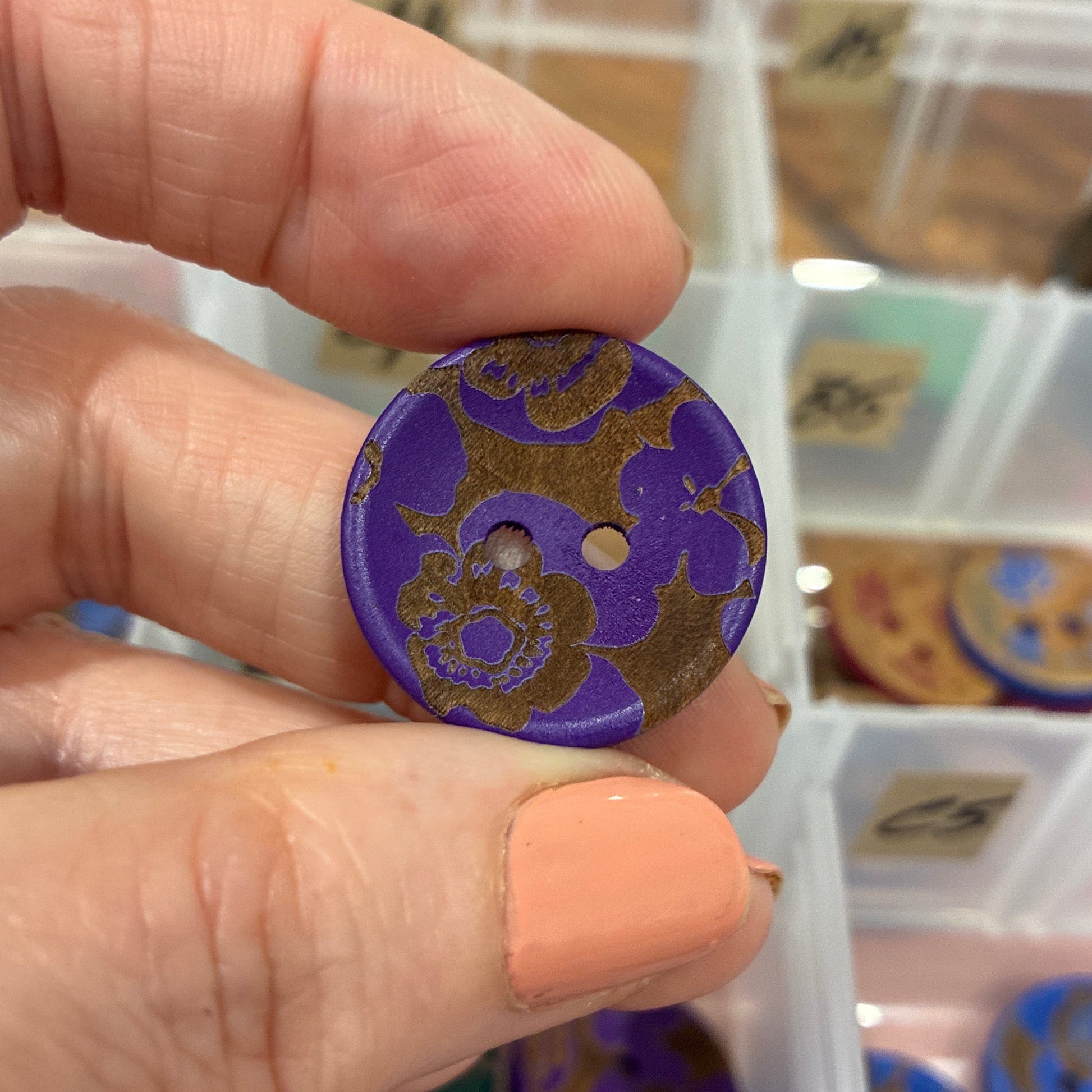 Hand holding a purple button with floral design in front of a container of buttons.