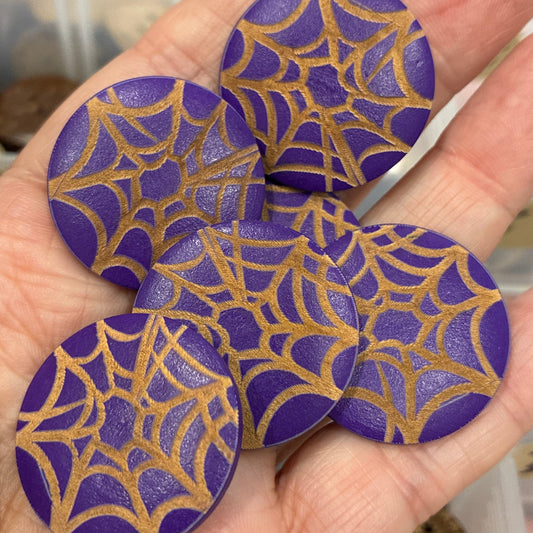 Hand holding three purple and gold spider web designs against a blurred background of buttons.