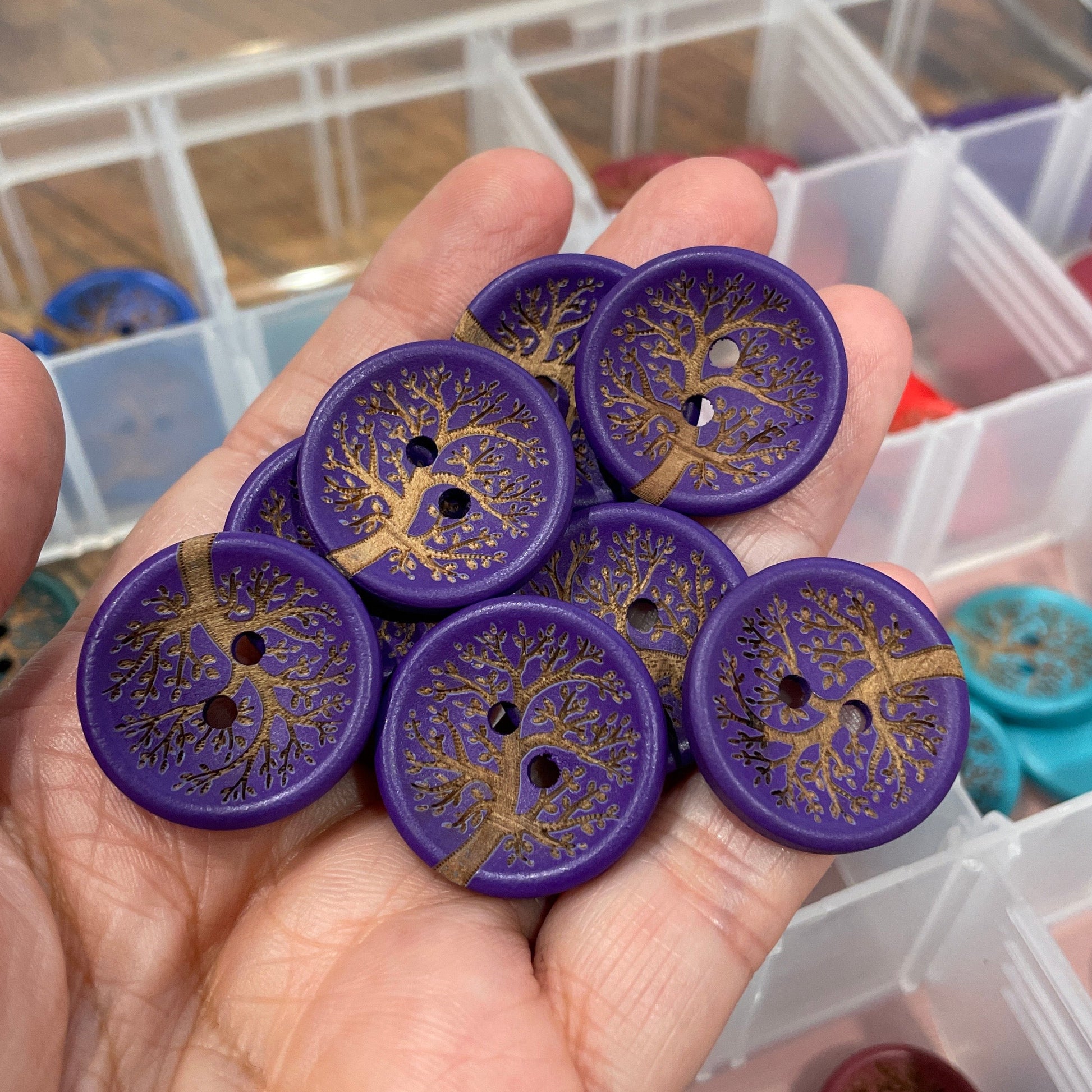 Hand holding purple buttons with gold designs, with a blurred background of containers.