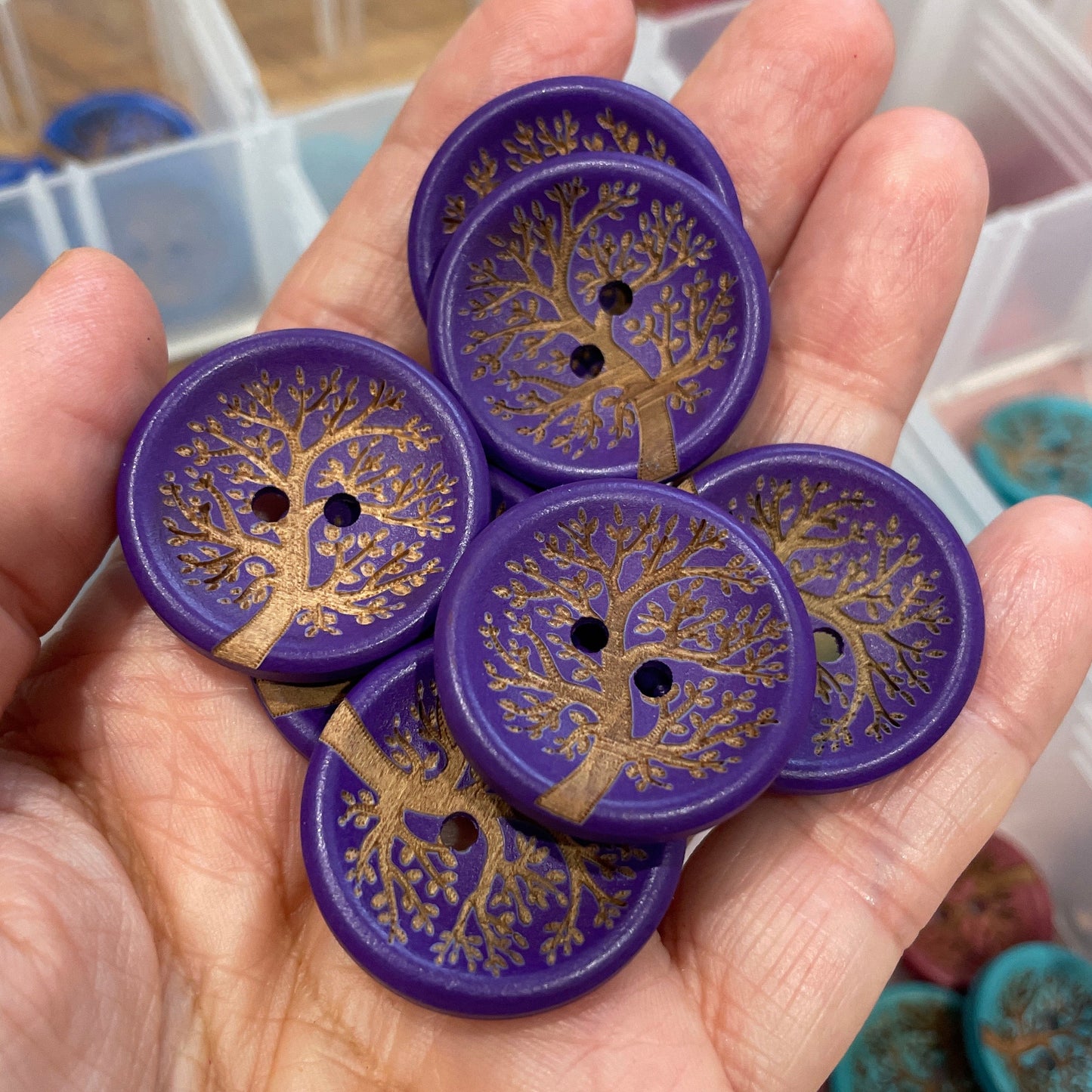 Hand holding purple buttons with tree design against a blurred background of a container.