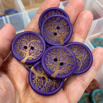 Hand holding purple buttons with tree design against a blurred background of a container.