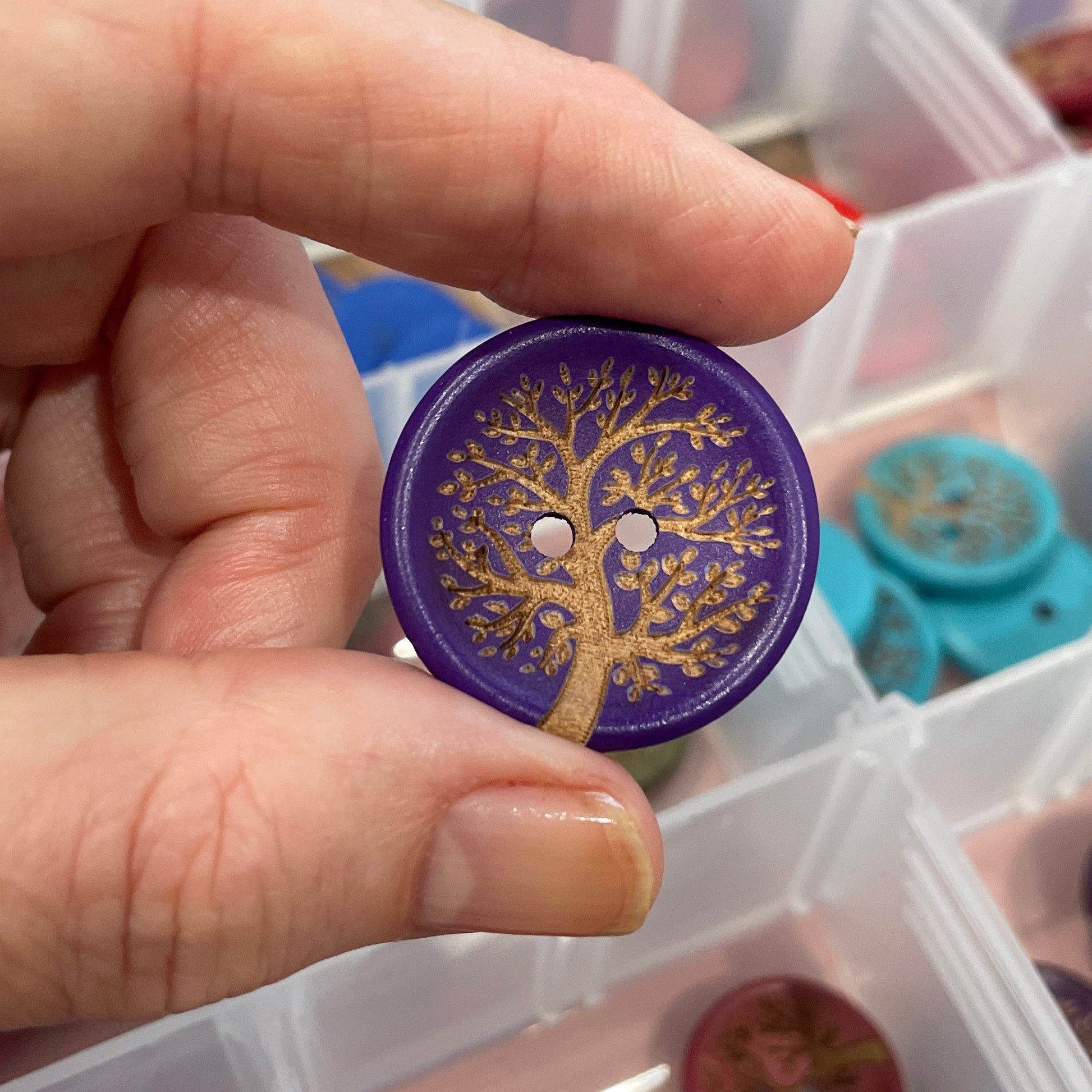 Hand holding a decorative button with intricate design, with a clear plastic container of buttons in the background.