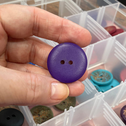 Hand holding a purple button with a container of assorted buttons in the background