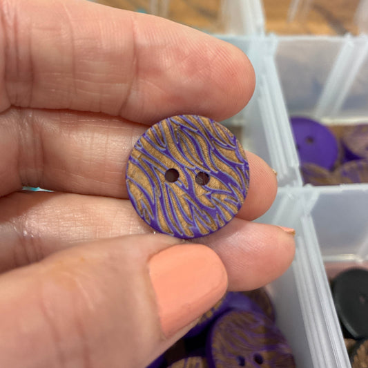 Hand holding a purple and gold button with a clear container of buttons in the background